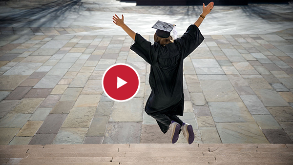 A woman in cap and gown leaping in front of Bailey Hall